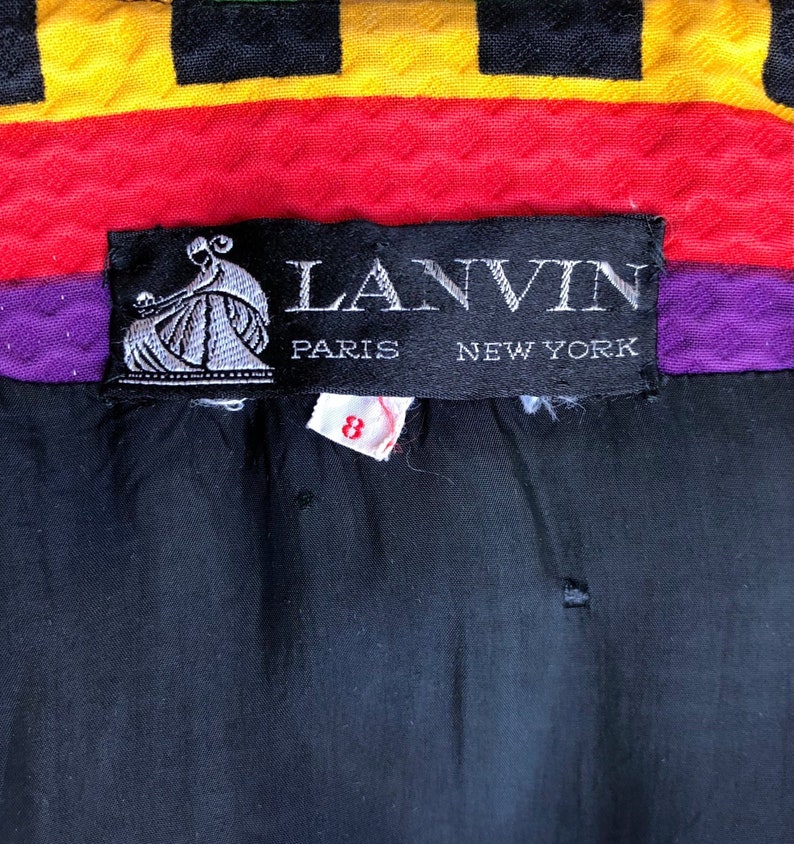 May include: A black garment label with white text reading LANVIN PARIS NEW YORK and a graphic of a person. Below the label is a small white tag with the red number 8. The fabric above the label features horizontal stripes of yellow, black, red, and purple.