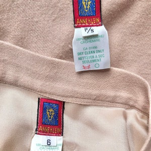 May include: Close-up of two sections of a light tan garment, possibly a skirt or trousers, with labels. The labels feature the "ANNE KLEIN" logo in red and blue, along with text indicating the fabric is 100% cashmere. One label indicates size P/S, the other size 6.