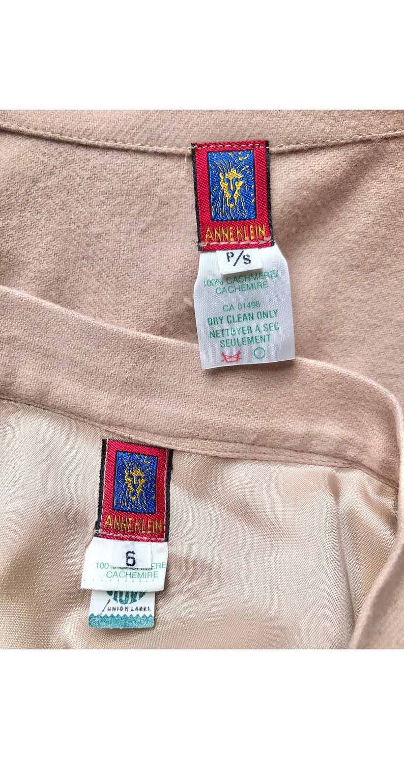 May include: Close-up of two sections of a light tan garment, possibly a skirt or trousers, with labels. The labels feature the "ANNE KLEIN" logo in red and blue, along with text indicating the fabric is 100% cashmere. One label indicates size P/S, the other size 6.