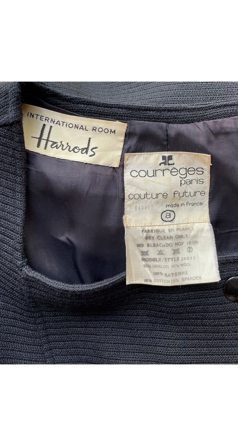 Op de afbeelding: Een zwarte blazer met een wit label met de tekst "International Room Harrods" en een wit label met de tekst "courr&egrave;ges paris couture future 040863 made in france" en wasinstructies.