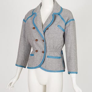 K&ouml;nnte beinhalten: Ein grauer Blazer mit blauen Zierleisten und Kn&ouml;pfen. Die doppelreihige Jacke hat eine taillierte Passform, dreiviertel &Auml;rmel und einen runden Kragen. Die blaue Zierleiste umrandet Kragen, Taschen und Manschetten. Die Jacke wird auf einer Schaufensterpuppe pr&auml;sentiert.