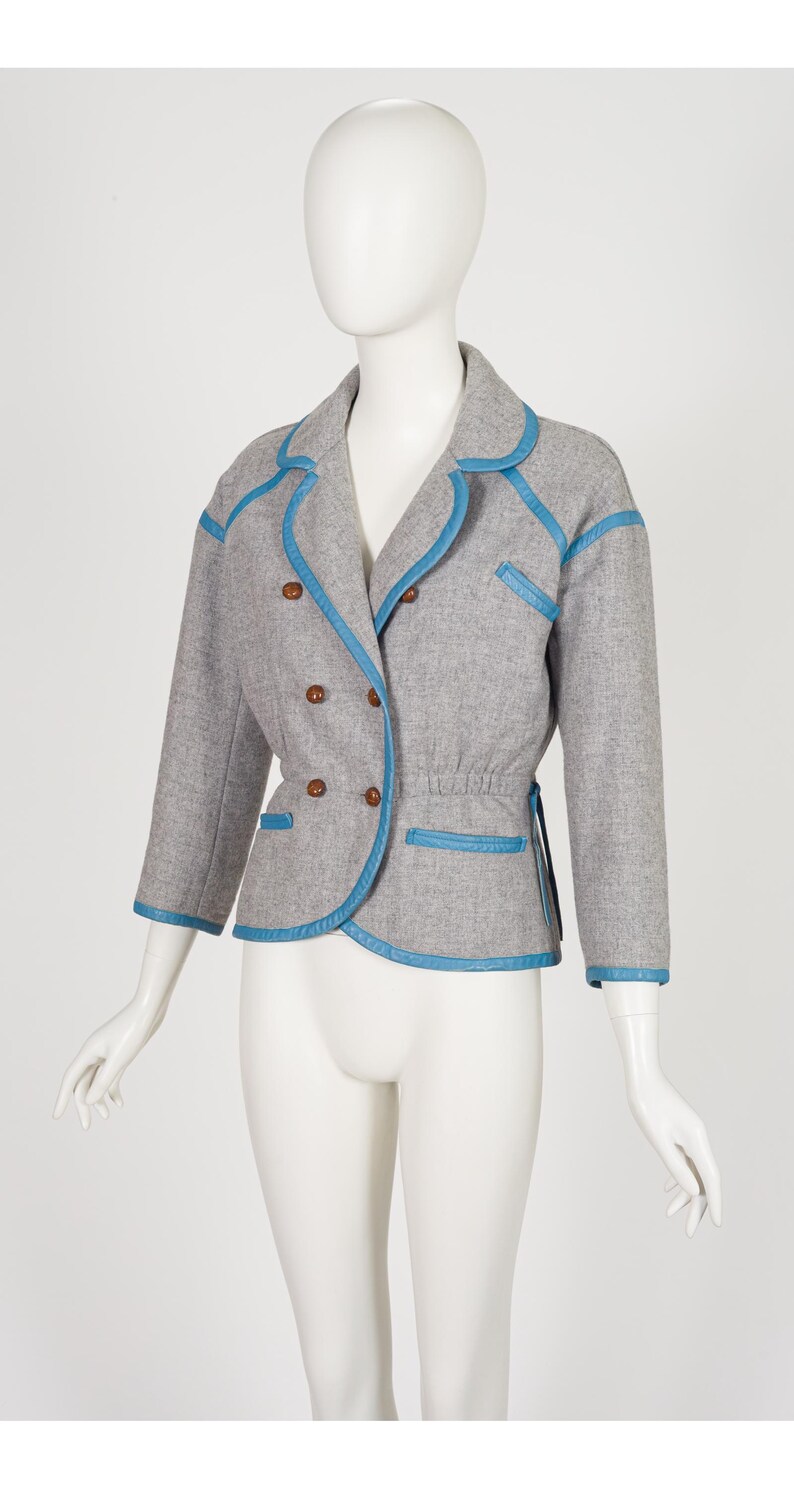 K&ouml;nnte beinhalten: Ein grauer Blazer mit blauen Zierleisten und Kn&ouml;pfen. Die doppelreihige Jacke hat eine taillierte Passform, dreiviertel &Auml;rmel und einen runden Kragen. Die blaue Zierleiste umrandet Kragen, Taschen und Manschetten. Die Jacke wird auf einer Schaufensterpuppe pr&auml;sentiert.
