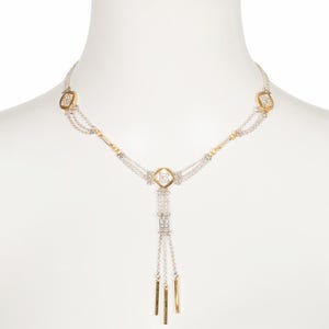 Puede incluir: Un collar plateado y dorado con un diseño geométrico. El collar presenta una serie de pequeños acentos cuadrados y rectangulares, con un colgante central de barras colgantes. El collar se muestra en un maniquí blanco.