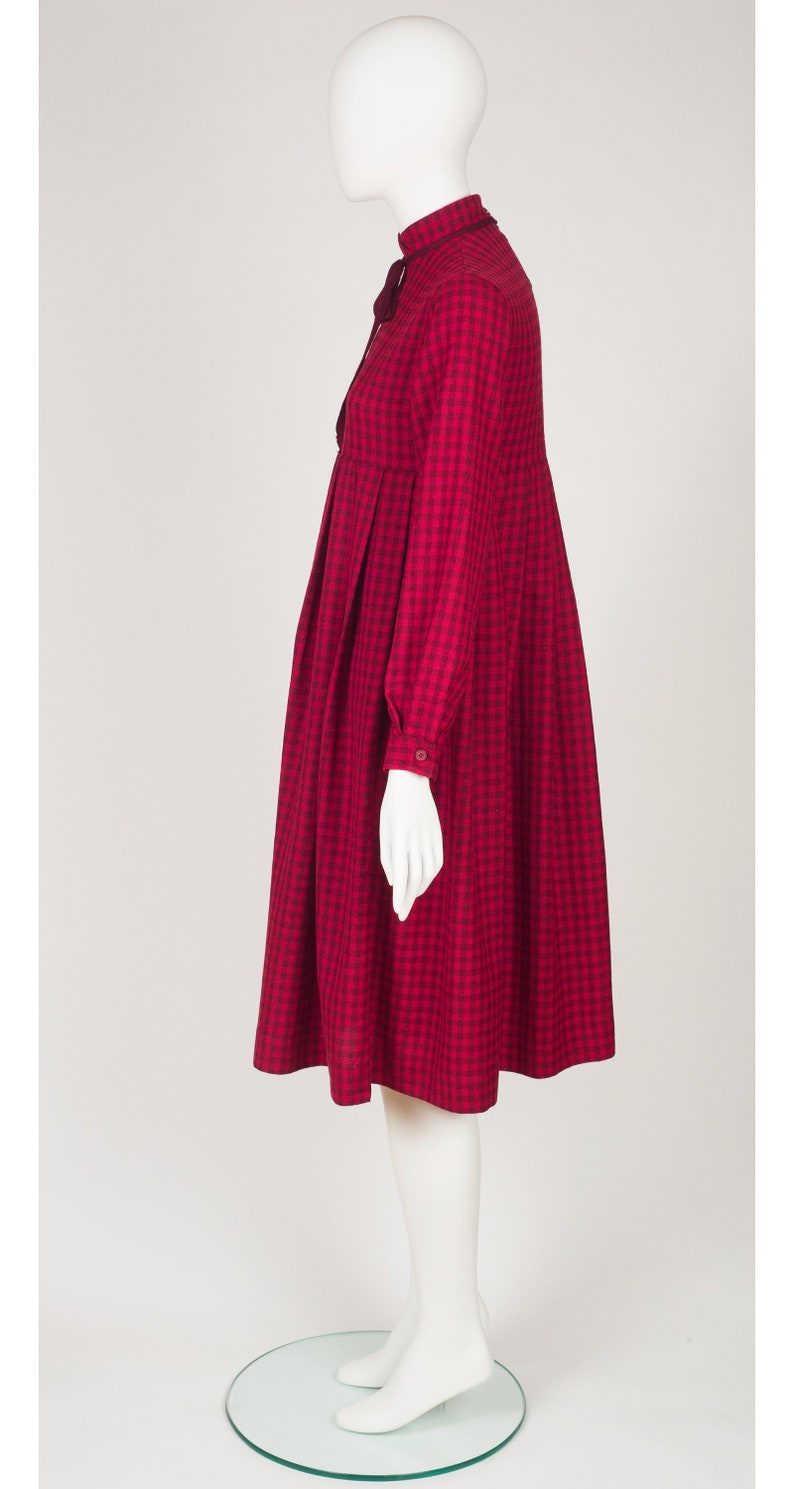 May include: A red and black gingham dress with a high neck and long sleeves. The dress has a gathered skirt and a bow at the neck.