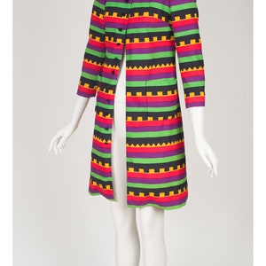 May include: A knee-length coat featuring a vibrant horizontal striped pattern in purple, green, black, and yellow. The coat has a collared neckline, three-quarter length sleeves, a front button closure, and two patch pockets on the front panels.