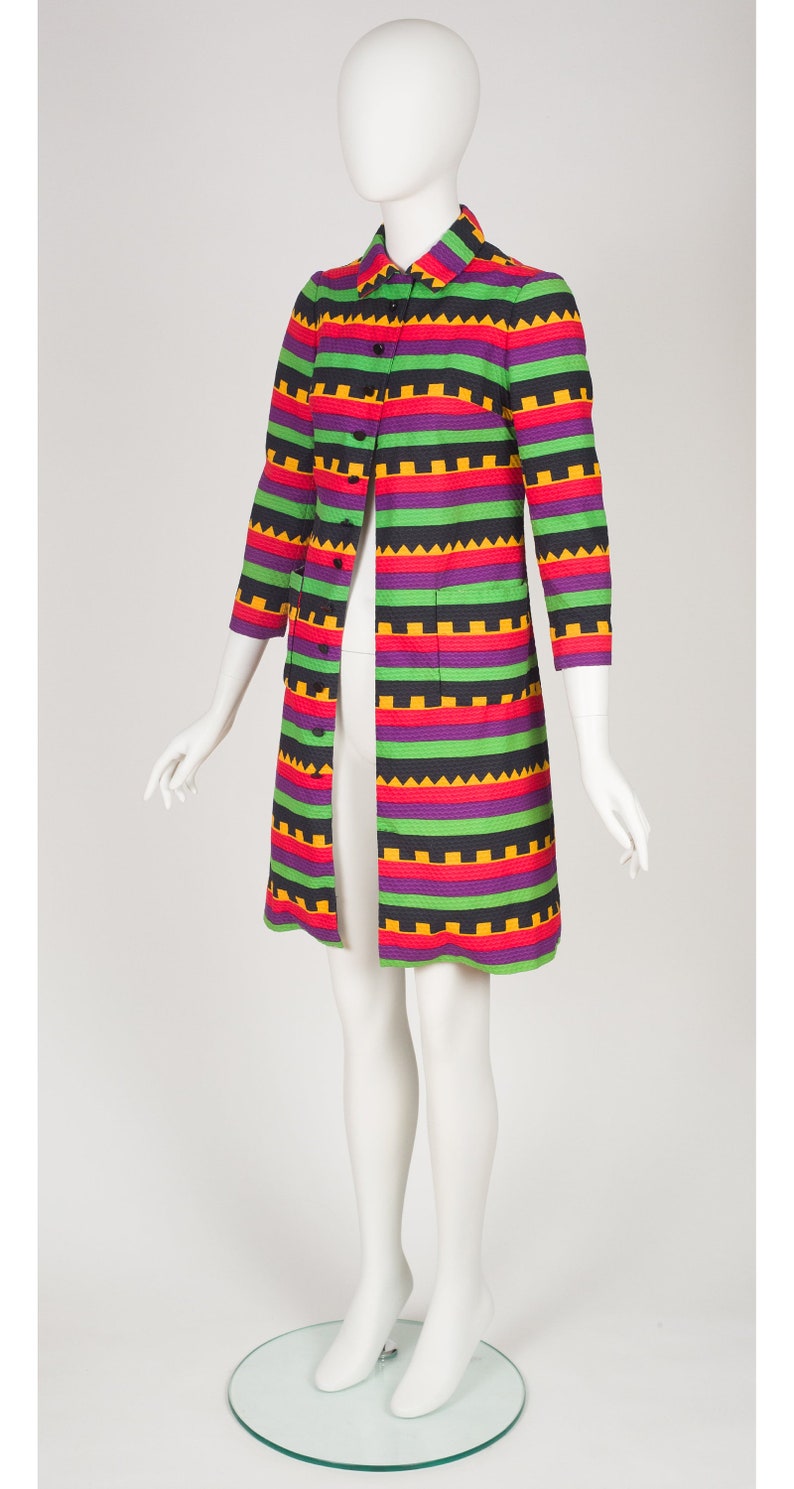 May include: A knee-length coat featuring a vibrant horizontal striped pattern in purple, green, black, and yellow. The coat has a collared neckline, three-quarter length sleeves, a front button closure, and two patch pockets on the front panels.
