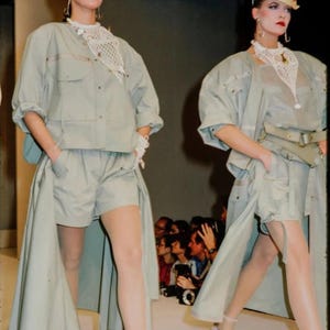 K&ouml;nnte beinhalten: Zwei Models auf einem Laufsteg tragen passende hellgr&uuml;ne Outfits. Jedes Outfit besteht aus einer kurz&auml;rmeligen Jacke, Shorts und einem langen, rockartigen Panel. Sie tragen H&uuml;te, geh&auml;kelte Halsb&auml;nder und Sandalen. Das Publikum ist im Hintergrund zu sehen.