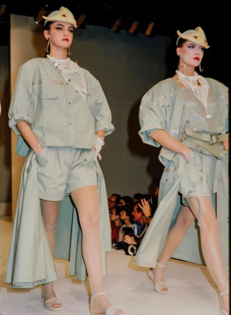 K&ouml;nnte beinhalten: Zwei Models auf einem Laufsteg tragen passende hellgr&uuml;ne Outfits. Jedes Outfit besteht aus einer kurz&auml;rmeligen Jacke, Shorts und einem langen, rockartigen Panel. Sie tragen H&uuml;te, geh&auml;kelte Halsb&auml;nder und Sandalen. Das Publikum ist im Hintergrund zu sehen.