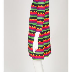 May include: A knee-length dress featuring a vibrant horizontal striped pattern in black, purple, green, red, and yellow. The garment has three-quarter length sleeves, a collared neckline, and a repeating geometric sawtooth design throughout.