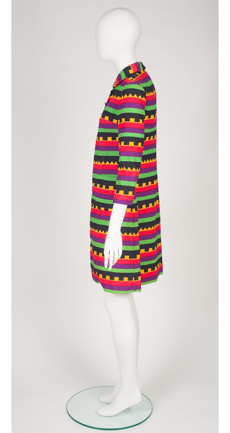 May include: A knee-length dress featuring a vibrant horizontal striped pattern in black, purple, green, red, and yellow. The garment has three-quarter length sleeves, a collared neckline, and a repeating geometric sawtooth design throughout.