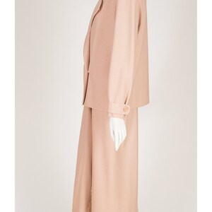 May include: A peach-coloured skirt suit on a mannequin. The suit features a blazer with shoulder pads and a knee-length skirt. The blazer has button details on the sleeves and a single button closure. The outfit is made of a smooth fabric.