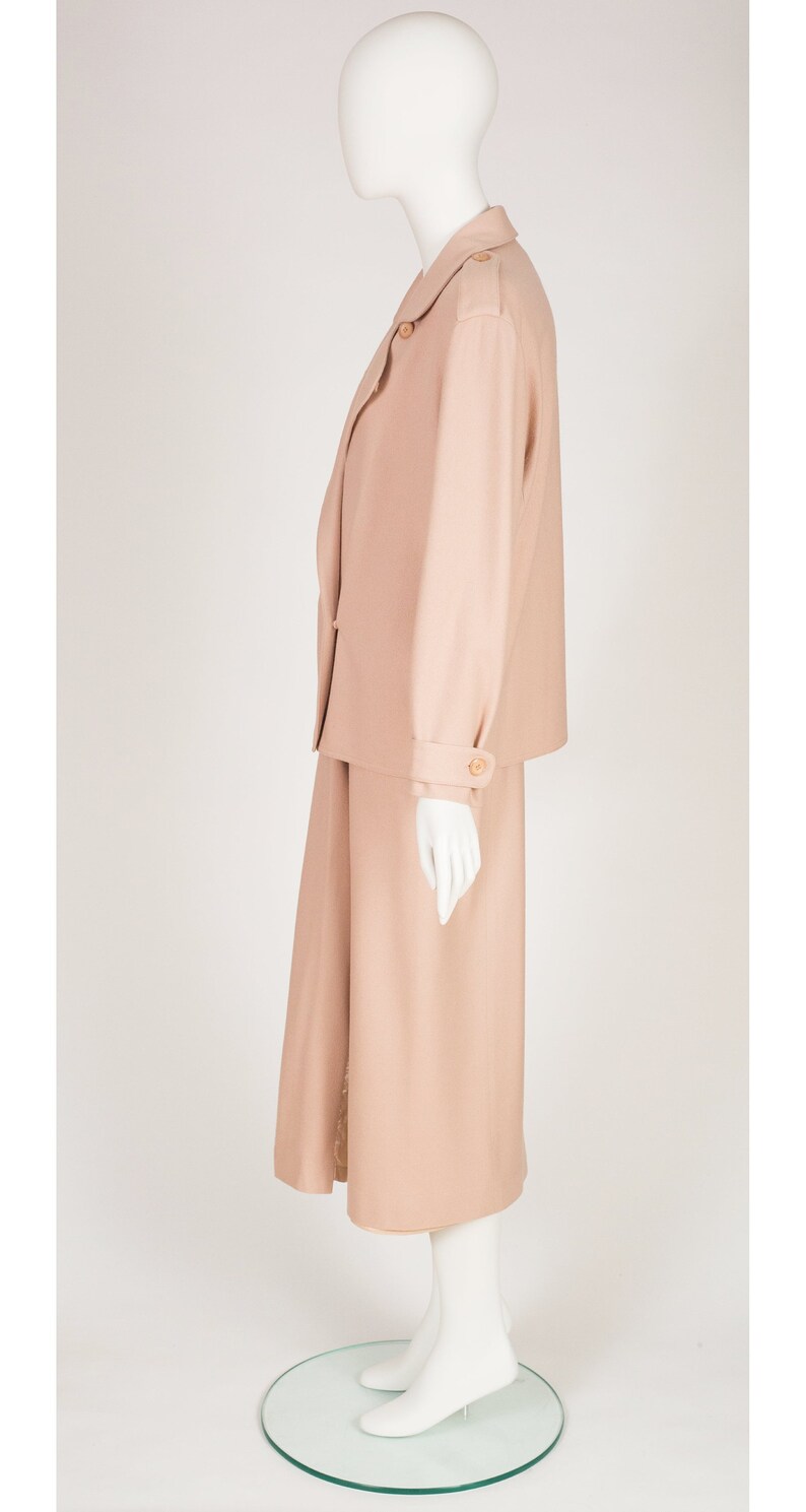 May include: A peach-coloured skirt suit on a mannequin. The suit features a blazer with shoulder pads and a knee-length skirt. The blazer has button details on the sleeves and a single button closure. The outfit is made of a smooth fabric.