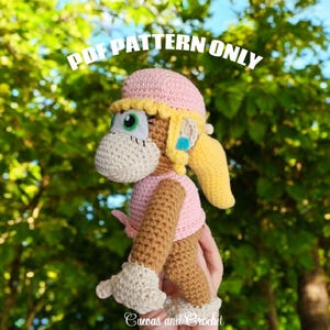 Może przedstawiać: Szydełkowa lalka małpki w kolorze różowym, żółtym i brązowym, z napisem "PDF PATTERN ONLY". Lalka ma różowy kapelusz, żółte włosy i różową koszulę. Lalka jest trzymana w dłoni osoby.