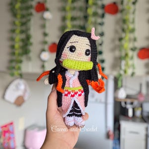 May include: Crocheted doll featuring a character with black hair, a pink kimono, and a green mouth guard. The doll has large eyes and is held in a hand. The doll is handmade and has orange accents. The text "Cuevas and Crochet" is visible.
