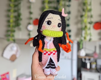 Op de afbeelding: Handgemaakte gehaakte pop van een personage met zwart haar, een roze kimono en een groene mondbeschermer. De pop heeft grote ogen en wordt in een hand gehouden. De pop heeft oranje accenten. De tekst "Cuevas and Crochet" is zichtbaar.