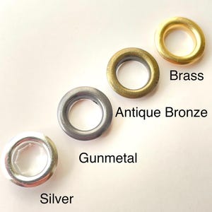 May include: Four metal grommets in brass, antique bronze, gunmetal, and silver. Each grommet is circular with a central hole. The names of the colors are printed below each grommet. These are used for crafting and sewing projects.