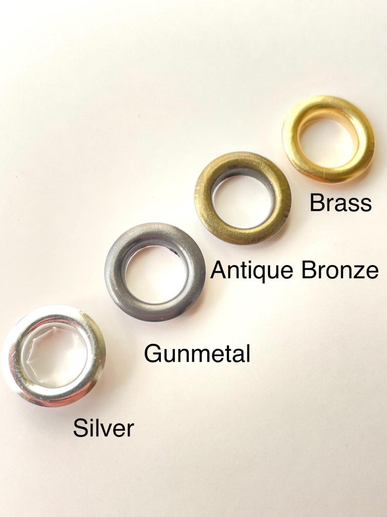 May include: Four metal grommets in brass, antique bronze, gunmetal, and silver. Each grommet is circular with a central hole. The names of the colors are printed below each grommet. These are used for crafting and sewing projects.