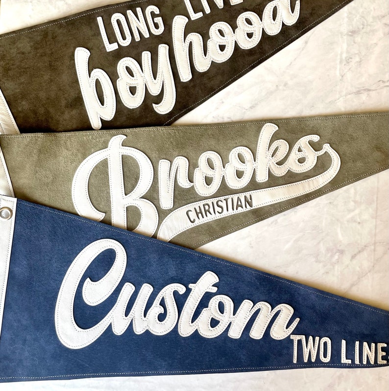 May include: Three felt pennants in shades of brown, green, and blue. Each pennant features white lettering. The top pennant reads "LONG LIVE boyhood", the middle one says "Brooks CHRISTIAN", and the bottom one says "Custom TWO LINE".