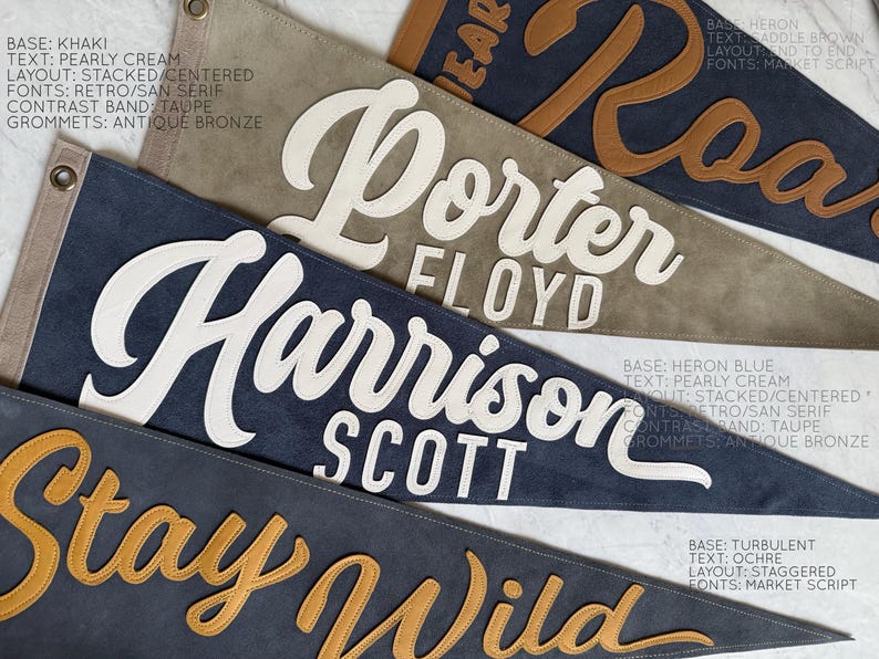 May include: Close-up of several personalized pennant flags in various colors, including khaki, heron blue, and turbulent. Each flag features custom text in contrasting colors and fonts, with antique bronze grommets.