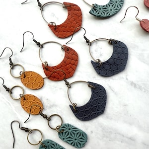 Puede incluir: Una colección de pendientes hechos a mano en varios colores y diseños. Los pendientes presentan cuero texturizado en tonos naranja, azul y verde azulado, con patrones geométricos y aros de metal. Algunos pendientes tienen formas ovaladas o de media luna.