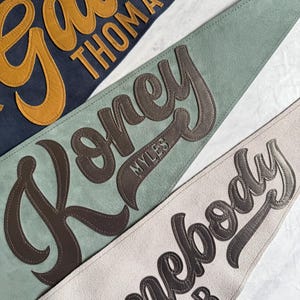 May include: Close-up of three personalized pennant flags in navy, sage green, and cream. Each pennant features a name in a contrasting color, with a stitched leather-like appearance. The pennants are likely made of felt or a similar material.