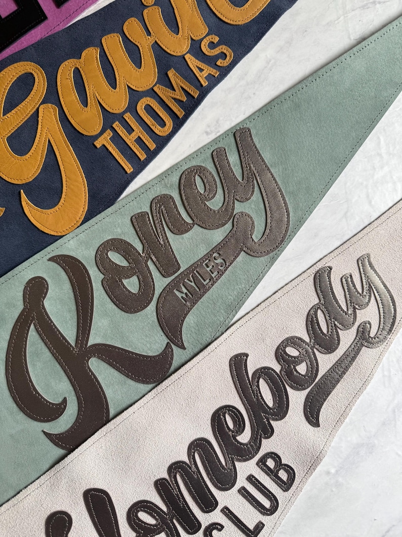 May include: Close-up of three personalized pennant flags in navy, sage green, and cream. Each pennant features a name in a contrasting color, with a stitched leather-like appearance. The pennants are likely made of felt or a similar material.