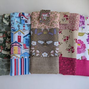 May include: Several fabric phone cases with diverse patterns. Designs feature cats, beach huts, floral prints, and a bee motif. Each case has a flap closure with a button or snap.