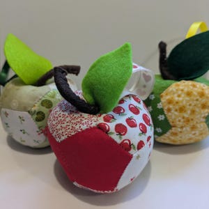 May include: Three fabric apple ornaments. Each apple is made from patchwork fabric in various patterns and colours, including red, white, green, and yellow. They have green felt leaves and brown stems, with ribbon accents.