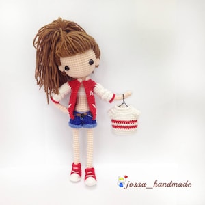 May include: Crocheted doll with brown hair wearing a red and white jacket, blue denim shorts, and red sneakers. The doll is holding a white and red striped crocheted shirt on a hanger.