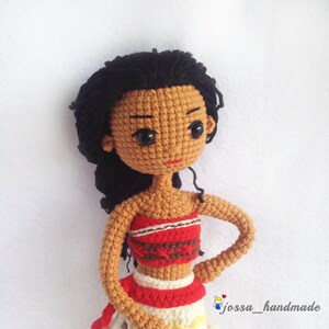May include: Crocheted doll with black hair, brown skin, and a red and white top. The doll is wearing a red and white skirt and has a red mouth.