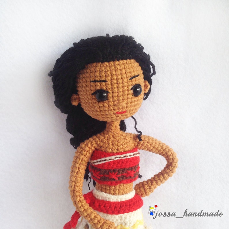 May include: Crocheted doll with black hair, brown skin, and a red and white top. The doll is wearing a red and white skirt and has a red mouth.