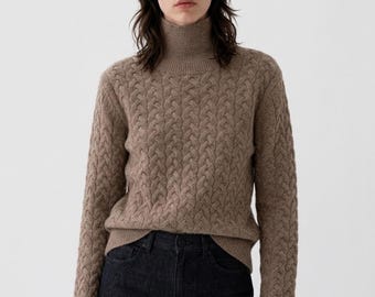 May include: A long-sleeve, light brown turtleneck sweater featuring a vertical cable knit pattern throughout the body and sleeves, paired with dark charcoal denim jeans. The sweater has a ribbed collar, cuffs, and hemline for a fitted look.
