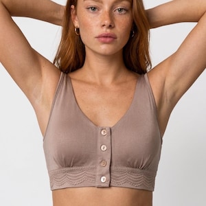 May include: A taupe-colored cropped tank top featuring a V-neckline, a front button placket with four light-colored buttons, and a decorative scalloped lace trim along the bottom hem. The garment is made of a smooth, solid-colored fabric.
