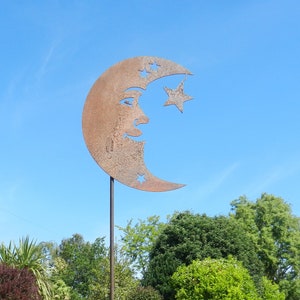 May include: A rusty metal crescent moon with a star and a face cut out, mounted on a tall metal stake. The moon is in a garden setting with green grass and trees in the background.