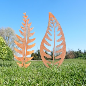 May include: Two rusty metal leaf sculptures in a grassy field. The leaves are cut out in a fern and a simple leaf shape.