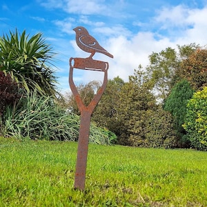 May include: A rusty metal garden stake with a bird silhouette perched on top. The stake is in a grassy area with green foliage in the background.
