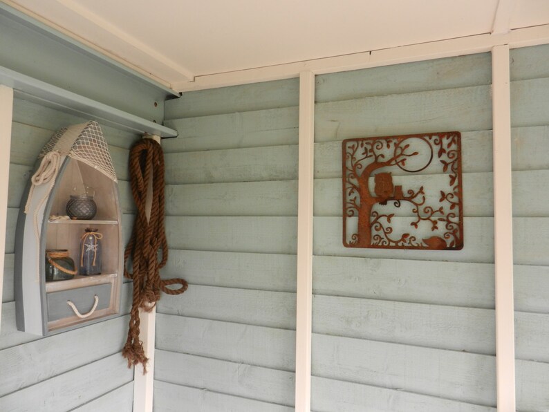 May include: A light blue wooden wall with a white trim. A small wooden boat shelf with a net and jars is on the left side of the wall. A rusty metal owl and tree wall decor is on the right side of the wall.