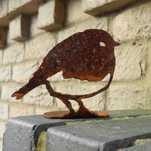 May include: A rusty metal bird sculpture perched on a branch. The bird has a simple, cutout design with a round body and a small beak. The sculpture is placed on a dark gray surface with a light-colored brick wall in the background.