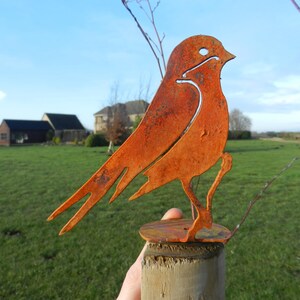 May include: A rusty orange metal bird sculpture. The bird is perched on a wooden post. The sculpture has a cutout design and is set against a background of green grass and a blue sky.