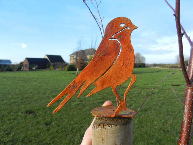 May include: A rusty orange metal bird sculpture. The bird is perched on a wooden post. The sculpture has a cutout design and is set against a background of green grass and a blue sky.