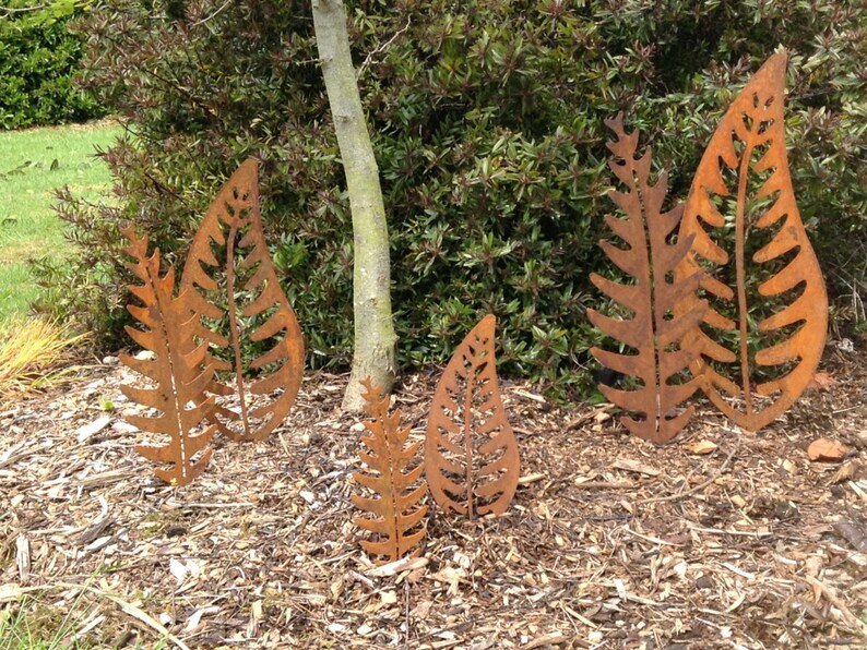 May include: Four rusty metal fern leaf sculptures of varying sizes are arranged in a garden setting. The sculptures are made of thin metal and have a rustic, weathered look.