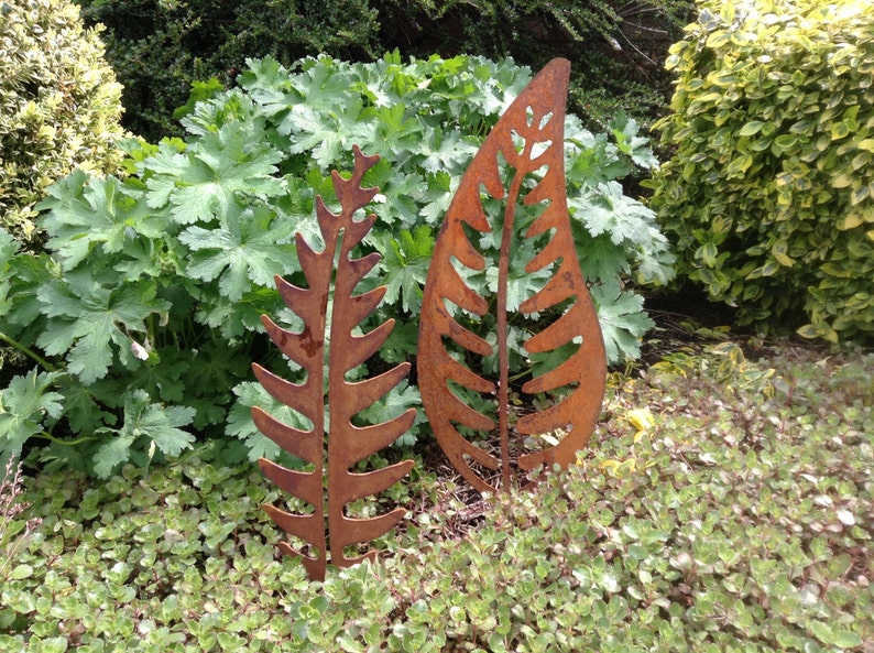 May include: Two rusty metal fern leaf sculptures in a garden setting. The larger leaf is taller than the smaller leaf.