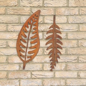 May include: Two rusty metal leaf-shaped wall decorations on a brick wall. The leaves are cut out and have a fern-like design.