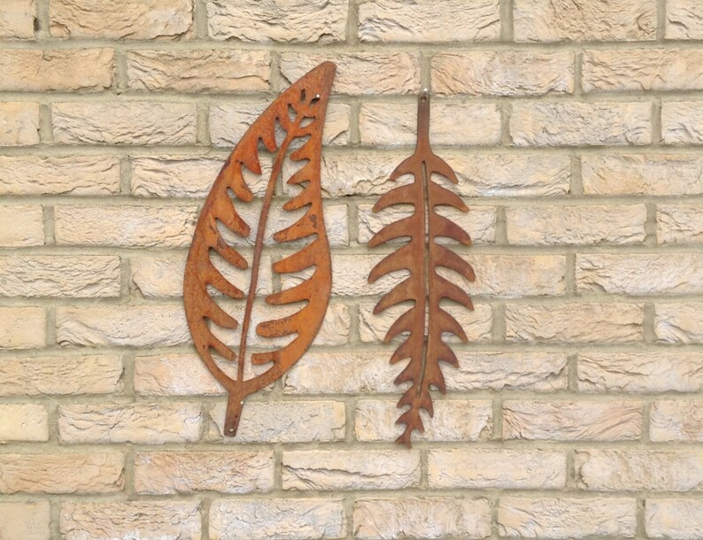 May include: Two rusty metal leaf-shaped wall decorations on a brick wall. The leaves are cut out and have a fern-like design.