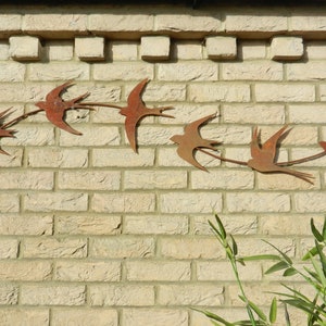 Op de afbeelding: Een bakstenen muur met een rij roestige metalen vogels in de vlucht. De vogels zijn uitsnijdingen en zijn met schroeven aan de muur bevestigd.