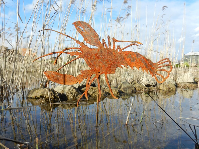 May include: A rusty metal lobster sculpture stands in a shallow pond with reeds in the background. The sculpture is a silhouette of a lobster with its claws outstretched.
