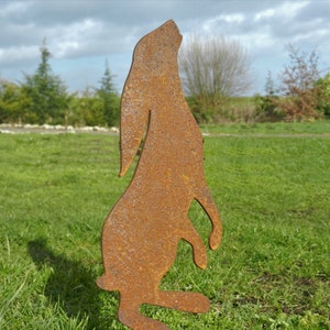 Puede incluir: Una silueta de metal oxidado de un conejo de pie sobre sus patas traseras, con la cabeza inclinada hacia arriba. La silueta está en un campo de hierba con un fondo borroso de árboles y cielo.