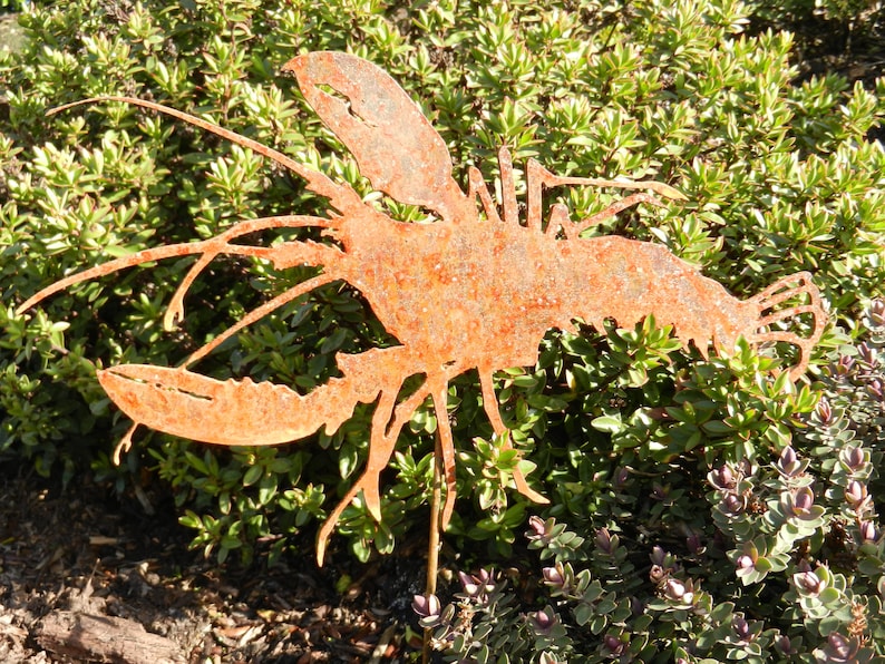 May include: A rusty metal lobster sculpture with a detailed design. The sculpture is standing upright in a garden setting.