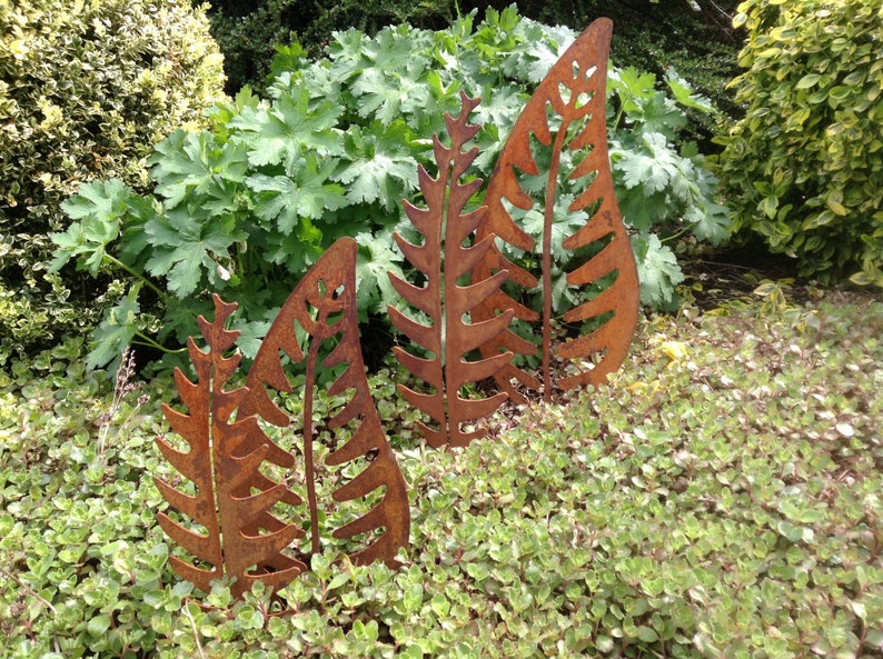May include: Three rusty metal fern leaf sculptures in a garden setting. The sculptures are different sizes and are arranged in a row.