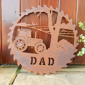Op de afbeelding: Een roestige metalen zaagblad met een silhouet van een tractor in een veld met bomen en het woord "DAD" uitgesneden.